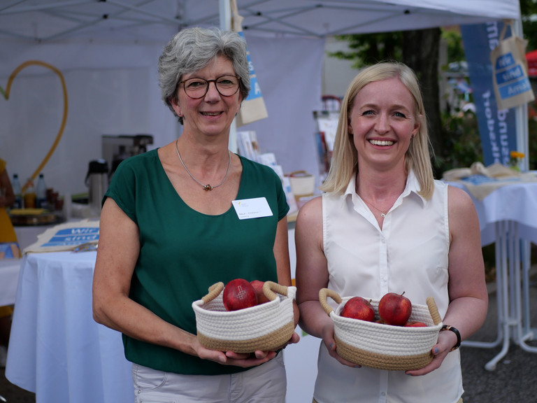 Zwei Mitarbeiterinnen des Amalie Sieveking Krankenhauses begrüßen Gäste mit Äpfeln am Stand des Gesundheitscampus Volksdorf beim Volksdorfer Stadtteilfest in Hamburg-Volksdorf