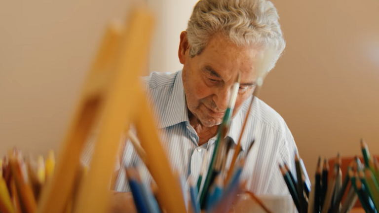 Ein Ehrenamtlicher der Kunsttherapie im Film Gesehen-Fühlen-Leben von Filmemacher Andrew Agbottah über die Menschen im Diakonie Hospiz Wannsee in Berlin sitzt an einem Tisch mit Kunst- und Malutensilien und blickt auf ein Bild Ein Ehrenamtlicher der Kunsttherapie im Film Gesehen-Fühlen-Leben von Filmemacher Andrew Agbottah über die Menschen im Diakonie Hospiz Wannsee in Berlin sitzt an einem Tisch mit Kunst- und Malutensilien und blickt auf ein Bild