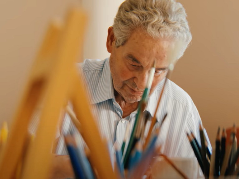 Ein Ehrenamtlicher der Kunsttherapie im Film Gesehen-Fühlen-Leben von Filmemacher Andrew Agbottah über die Menschen im Diakonie Hospiz Wannsee in Berlin sitzt an einem Tisch mit Kunst- und Malutensilien und blickt auf ein Bild Ein Ehrenamtlicher der Kunsttherapie im Film Gesehen-Fühlen-Leben von Filmemacher Andrew Agbottah über die Menschen im Diakonie Hospiz Wannsee in Berlin sitzt an einem Tisch mit Kunst- und Malutensilien und blickt auf ein Bild