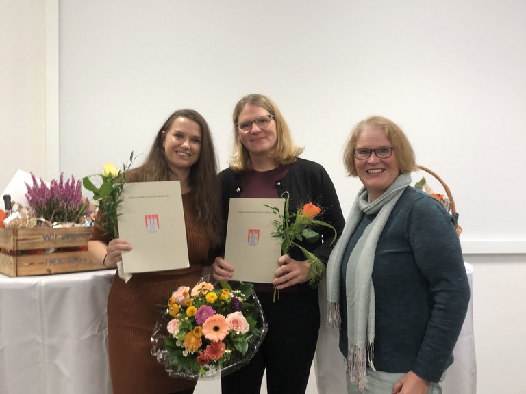 Drei Frauen stehen bei einer Abschlussfeier zusammen; zwei von ihnen halten Zertifikate und Blumensträuße nach bestandener Weiterbildung zur Hygienefachkraft. Drei Frauen stehen bei einer Abschlussfeier zusammen; zwei von ihnen halten Zertifikate und Blumensträuße nach bestandener Weiterbildung zur Hygienefachkraft.