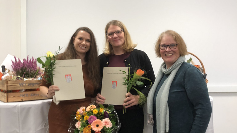 Drei Frauen stehen bei einer Abschlussfeier zusammen; zwei von ihnen halten Zertifikate und Blumensträuße nach bestandener Weiterbildung zur Hygienefachkraft. Drei Frauen stehen bei einer Abschlussfeier zusammen; zwei von ihnen halten Zertifikate und Blumensträuße nach bestandener Weiterbildung zur Hygienefachkraft.