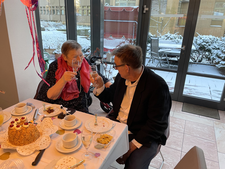 Das Hochzeitspaar sitzt am Kaffee-Tisch und stößt mit einem Glas Sekt an, besonderer Momente im Hospiz, Lebensabschnitt feiern, Diakonie Hospiz Wannsee, Berlin-Wannsee