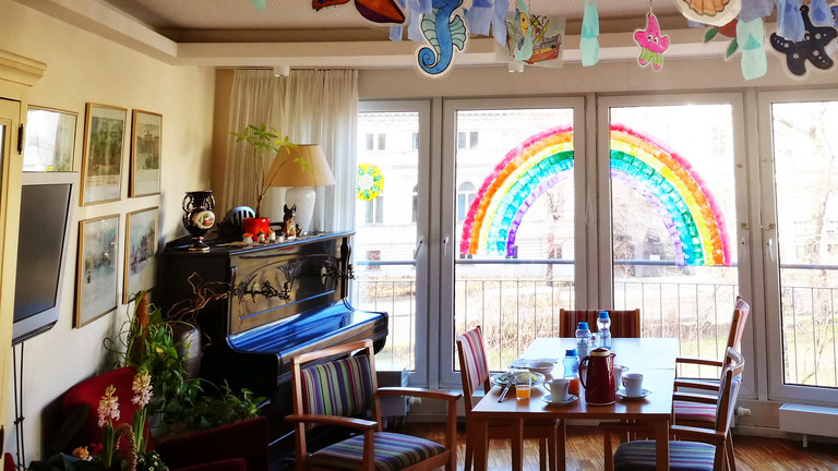 Wohnzimmer mit Regenbogen und Klavier, Queersensible Pflege, LGBTQ Berlin, Lesetipp Süddeutsche Zeitung, Endlich bei sich, Immanuel Seniorenzentrum Schöneberg Ein Wohnzimmer mit Regenbogen und Klavier im Immanuel Seniorenzentrum Schöneberg
