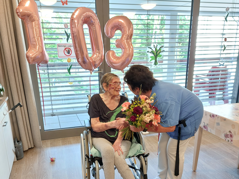 Bewohnerin im Rollstuhl bekommt Blumen von Pflegerin überreicht