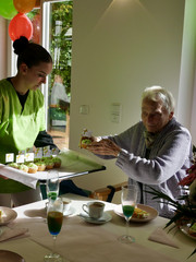 Eine Beschäftigte reicht einer am Tisch sitzenden Frau ein Tablett mit bunten Muffins bei der Wiedereröffnung der Tagespflegestätte für ältere Menschen im Immanuel Seniorenzentrum Kläre Weist in Petershagen Eine Beschäftigte reicht einer am Tisch sitzenden Frau ein Tablett mit bunten Muffins bei der Wiedereröffnung der Tagespflegestätte für ältere Menschen im Immanuel Seniorenzentrum Kläre Weist in Petershagen